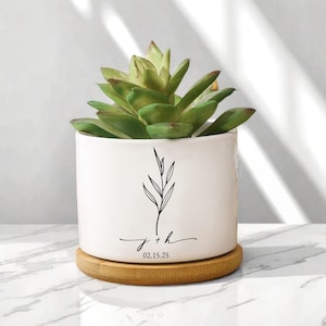 May include: A white ceramic plant pot with a green succulent. The pot features a black floral design, initials "j + h", and the date "02.15.25". It rests on a round, light brown wooden coaster. A decorative item.