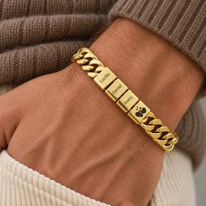 May include: A gold-toned cuban link chain bracelet featuring three rectangular metal beads engraved with the names Lucilla, Daniel, and Avery, followed by a fourth bead displaying a small graphic of three stacked hearts in black ink.