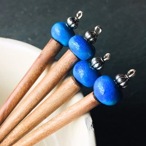 Puede incluir: Cuatro palitos de madera para el pelo con cuentas azules y plateadas en la parte superior. Los palitos están dispuestos en fila, con las cuentas mirando hacia la cámara.