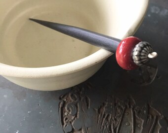 May include: A black wooden hair stick with a red and silver bead at the end. The stick is resting in a white ceramic bowl.