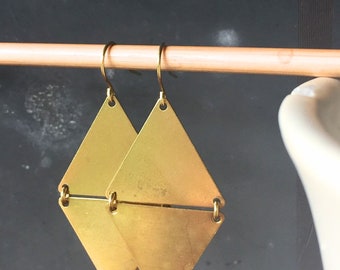 May include: A pair of gold-colored, diamond-shaped earrings. Each earring is a flat, geometric shape with a horizontal bar connecting the two halves. The earrings hang from gold-colored ear wires.
