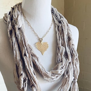 May include: A gold-colored leaf pendant necklace with a delicate chain, displayed on a mannequin. The necklace is paired with a long, light gray scarf with a textured, ribbon-like design. The scarf is draped around the mannequin's neck.