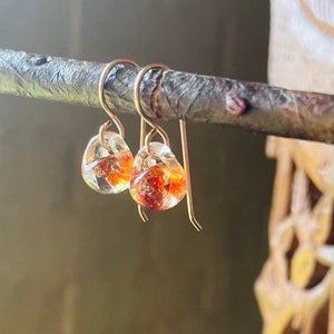 Op de afbeelding: Een paar gouden oorhangers met heldere en oranje glazen kralen. De kralen hebben een gevlekt, gemarmerd uiterlijk.