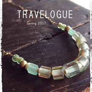 Könnte beinhalten: Eine grüne und goldene Halskette mit verschiedenen Perlen, darunter klare Glasperlen mit Streifen, grüne Glasperlen und goldene Perlen. Die Halskette liegt auf einer Holzoberfläche und der Text "TRAVELOGUE Frühling 2017" ist über der Halskette sichtbar.