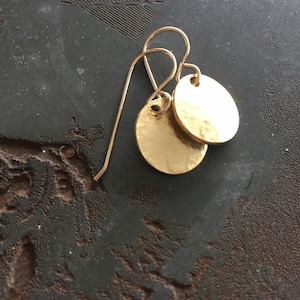 Puede incluir: Pendientes colgantes dorados con dos colgantes circulares martillados. Los pendientes tienen un diseño de gancho simple. Las joyas se exhiben sobre una superficie oscura y texturizada.