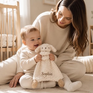 May include: A cream-colored plush security blanket featuring a stuffed animal head with floppy ears and a soft, textured body. The fabric is embroidered with the name Sophia in a dark brown script font, centered on the lower portion of the blanket.