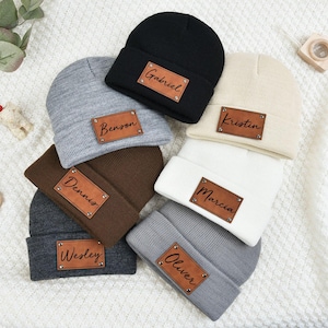 May include: Six gray, brown, black, and white knit beanies with leather patches that have names written on them. The names are Gabriel, Benson, Dennis, Marcia, Wesley, and Oliver.