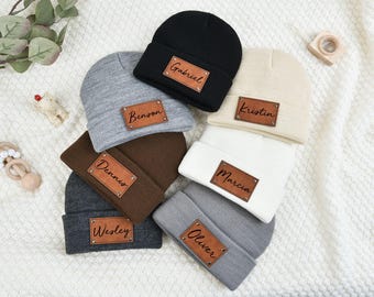 May include: Six gray, brown, black, and white knit beanies with leather patches that have names written on them. The names are Gabriel, Benson, Dennis, Marcia, Wesley, and Oliver.