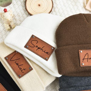 Pode incluir: Três gorros de malha com patches de couro personalizados. Os gorros são de cor castanho, branco e bege. Os patches têm os nomes "Sophia", "Austin" e "Violet" escritos neles.