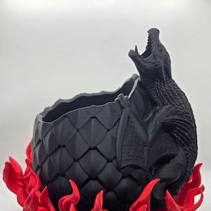 May include: A black dragon-themed planter with a textured egg-like body and a red flame base. The dragon is perched on the side with its mouth open. A unique decorative item for fantasy enthusiasts.