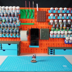 Op de afbeelding: Een oranje pegboard-organizer met blauwe en groene accenten, met verfpotten, penselen en andere knutselbenodigdheden. Inclusief lades, een telefoonhouder en een snijmat. Een miniatuurfiguur ligt op de mat.