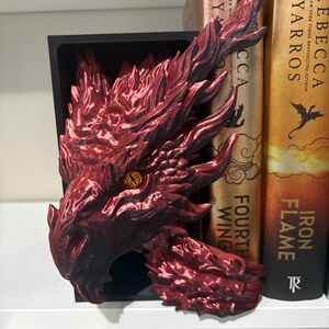 May include: A red dragon head bookend with a black base. The dragon has detailed scales and an open mouth, with a yellow eye. The bookend is placed on a white shelf next to books with titles like "Fourth Wing" and "Iron Flame".