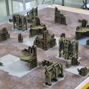 Peut inclure: Une collection de ruines de bâtiments miniatures de style gothique marron disposées sur un tapis de jeu texturé marron et gris. L'ensemble comprend diverses sections de murs, des pièces d'angle et des structures arquées détaillées.