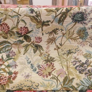 May include: Close-up of a floral tapestry with a cream background. The tapestry features a detailed pattern of various flowers in shades of pink, green, blue, and brown. The design includes leaves and stems, creating a natural, botanical theme.