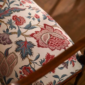 May include: Close-up of a chair with a floral pattern. The fabric features large red and purple flowers with blue leaves on a cream background. The wooden armrest is visible in the foreground.
