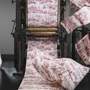Puede incluir: Una prensa de impresión antigua muestra rollos de tela con un diseño toile de Jouy rojo y blanco. La tela está drapeada y fluyendo, con una cortina a la izquierda y rollos de tela que pasan por la prensa. La escena evoca la historia y la artesanía.