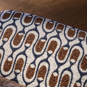 Op de afbeelding: Close-up van een patroonstof met een herhalend ontwerp. Het patroon heeft bruine, marineblauwe en gebroken witte kleuren. De stof lijkt bekledingsmateriaal te zijn, mogelijk voor een stoel of een poef.