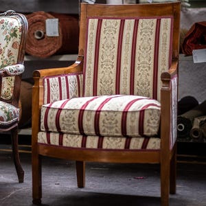 Op de afbeelding: Antieke houten fauteuil met een gestreepte rood-crèmekleurige rugleuning en zitkussen. De stoel heeft een lichtbruin houten frame en armleuningen. Een andere stoel met een bloemmotief is gedeeltelijk zichtbaar aan de linkerkant.