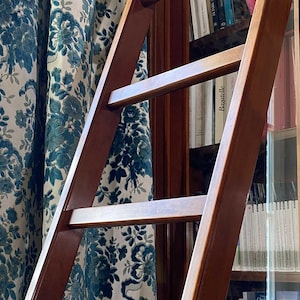 Op de afbeelding: Een houten bibliotheekladder met een donkerbruine afwerking leunt tegen een boekenkast. De treden van de ladder zijn zichtbaar en de achtergrond omvat een blauw-wit gordijn met patroon en een boekenplank.