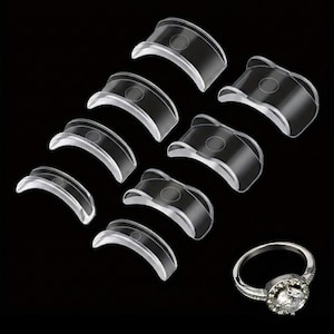 May include: Clear, curved plastic lash lift shields in various sizes are arranged with a silver ring featuring a large, round, clear gemstone. The shields are designed to lift and curl eyelashes. The background is black.