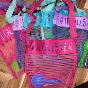 May include: Four pink mesh bags with colorful straps and the text "It Calls Me" printed on the front. Each bag contains various colorful plastic beach toys.