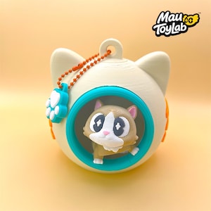 May include: A cream-colored spherical cat-shaped container with a teal circular frame opening. Inside sits a small brown and white cat figurine. An orange ball chain with a teal paw print charm is attached to the top. The text Mau ToyLab is visible.
