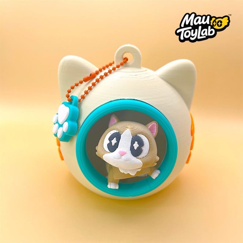 May include: A cream-colored spherical cat-shaped container with a teal circular frame opening. Inside sits a small brown and white cat figurine. An orange ball chain with a teal paw print charm is attached to the top. The text Mau ToyLab is visible.