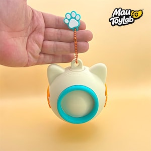 May include: A cream-colored, spherical cat-shaped toy with a teal circular center and orange side accents. It features a small loop at the top attached to an orange ball chain and a teal and white paw print charm. The text Mau ToyLab is visible.