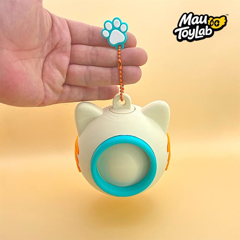 May include: A cream-colored, spherical cat-shaped toy with a teal circular center and orange side accents. It features a small loop at the top attached to an orange ball chain and a teal and white paw print charm. The text Mau ToyLab is visible.