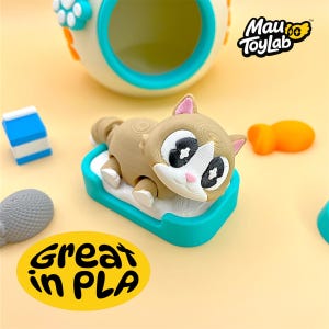 May include: A tan and white cat figurine with large black eyes and a pink nose, resting on a teal and white base. Nearby are a blue and white block, a gray textured object, and an orange shape. The text Mau ToyLab and Great in PLA appear.