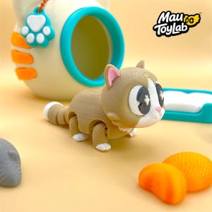 May include: A tan and white articulated cat figurine with large dark eyes and white paws, positioned in front of a cream-colored cat-shaped container with a teal rim and a paw-shaped tag. The Mau ToyLab logo is visible in the upper right corner.