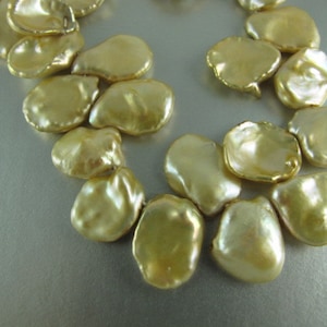 May include: A close-up view of a necklace featuring large, irregularly shaped, golden-colored pearls. The pearls have a lustrous, iridescent sheen, and are strung together to form a necklace. The background is a light, neutral color.