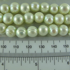 May include: A strand of off-white round pearls with a slight green hue. The pearls are approximately 1/2 inch in diameter and are measured against a ruler.