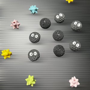 May include: A collection of small, round, textured black beads with white eyes, interspersed with multi-colored, star-shaped beads in shades of yellow, light blue, and pale pink, arranged in a scattered pattern against a dark gray background.