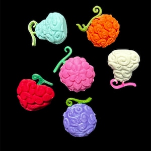 May include: A collection of six colorful, textured resin fruits, each with a unique shape and color. The fruits include shades of blue, orange, pink, white, red, and purple, with green stems. The background is solid black.