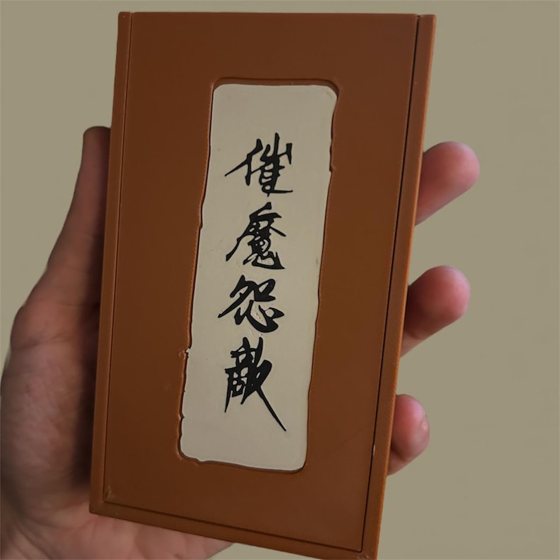 May include: A brown rectangular box with a white rectangular inset. The inset features black Japanese characters. The box is held in a hand.
