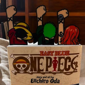 May include: A One Piece manga book with a skull and crossbones logo and the text "East Blue" and "One Piece" on the cover. Four character figurines are standing on top of the book, each with a raised fist and a cross on their arm.