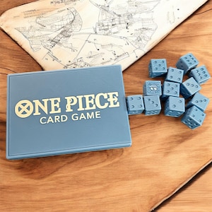 May include: A light blue "ONE PIECE CARD GAME" box with gold lettering and a set of light blue dice. The dice have white markings and a skull and crossbones symbol. A map is in the background.