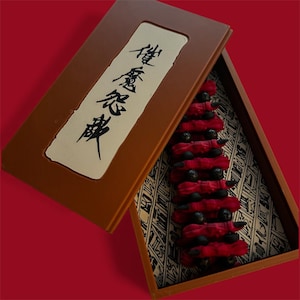 May include: A brown box with a hinged lid, containing a series of red, fabric-wrapped objects with black spheres. The box has a cream-colored label with black Japanese characters. The interior of the box has a patterned fabric.