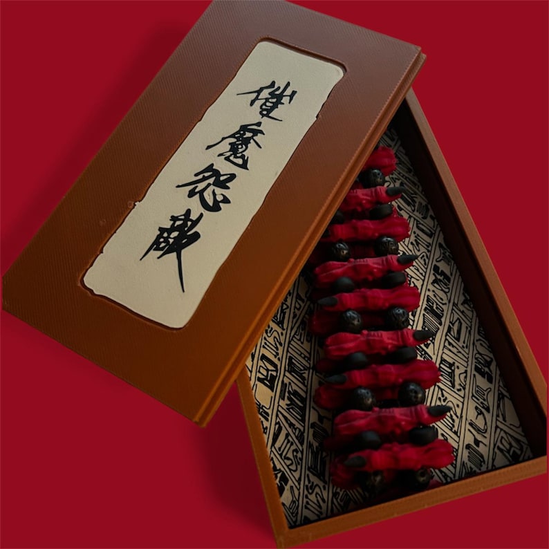 May include: A brown box with a hinged lid, containing a series of red, fabric-wrapped objects with black spheres. The box has a cream-colored label with black Japanese characters. The interior of the box has a patterned fabric.
