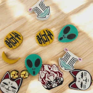 May include: A collection of assorted pins including two white and black patterned shapes with pink accents, two yellow circular pins with black text, two green alien head pins, a yellow banana, two white cat face pins, and one red and white anime character pin.