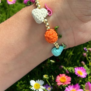 May include: A colorful beaded bracelet featuring a pink strawberry, white heart, orange sphere, and blue whale charm. The bracelet is strung with gold beads and accented with green and pink details. A unique and whimsical accessory.