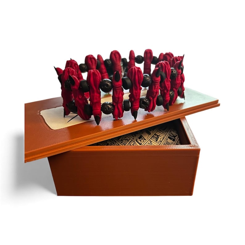 May include: A red and black beaded bracelet rests on an open, brown wooden box. The bracelet features red fabric-covered beads and black beads. The box has a decorative interior and a hinged lid.