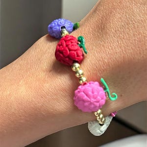 May include: A beaded bracelet featuring three colorful, brain-shaped charms in purple, red, and pink. The charms are connected by gold-colored beads and small green accents. A silver-colored charm hangs at the end.