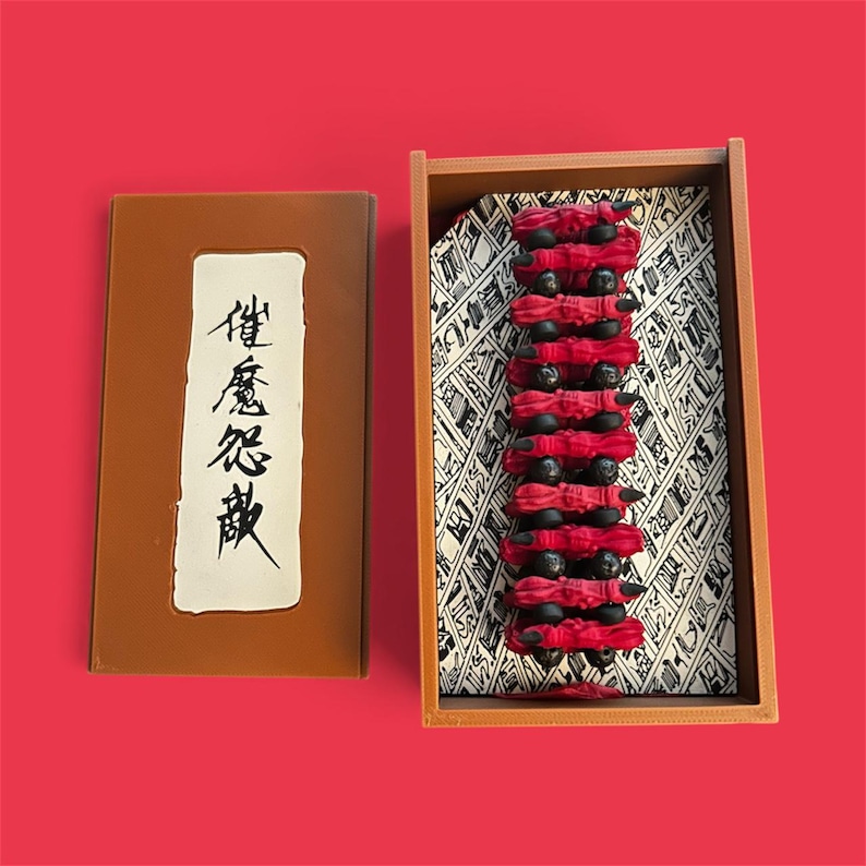 May include: A wooden box with a brown cover and a rectangular opening, next to a wooden tray. The tray contains a stack of red fabric pieces with black spheres. The cover has Japanese text on a white background.