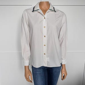 May include: White button-down shirt with long sleeves and a collared neckline. The collar is trimmed with a navy blue and green stripe. The shirt has gold-coloured buttons and is paired with blue jeans.