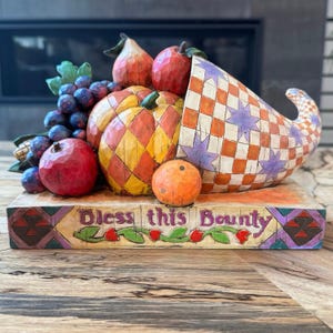 May include: A decorative cornucopia figurine featuring a checkered pattern with purple stars, overflowing with carved-style fruit including a pumpkin, grapes, and apples. The base is inscribed with the text Bless this Bounty in purple lettering.