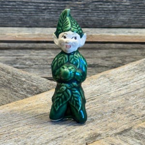 May include: A ceramic figurine of a green elf with a leaf-shaped hat and clothing. The elf has a white face with rosy cheeks and a small, smiling mouth. The figurine is posed in a kneeling position.