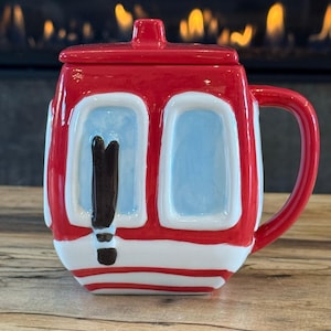 Op de afbeelding: Een rood-witte keramische mok in de vorm van een skigondel met een bijpassend deksel. De mok heeft lichtblauwe ramen en een zwarte skigrafiek op de voorzijde. Het heeft een stevig handvat aan de rechterkant en een platte onderkant.