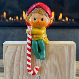 May include: A decorative elf figurine with a red hat, yellow shirt, and green pants, holding a red and white striped candy cane. The elf is designed to sit on the edge of a flat surface, featuring a smooth finish and painted facial details.